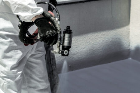 Facharbeiter trägt mit einem Sprühgerät eine Schutzbeschichtung auf eine Wandoberfläche auf, trägt weißen Schutzanzug und schwarze Handschuhe.