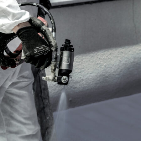 Facharbeiter trägt mit einem Sprühgerät eine Schutzbeschichtung auf eine Wandoberfläche auf, trägt weißen Schutzanzug und schwarze Handschuhe.
