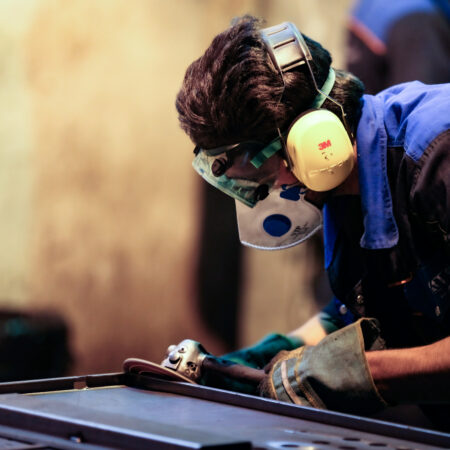 Arbeiter bearbeitet eine Metalloberfläche mit einem Schleifwerkzeug, trägt Schutzbrille, Maske und Gehörschutz.